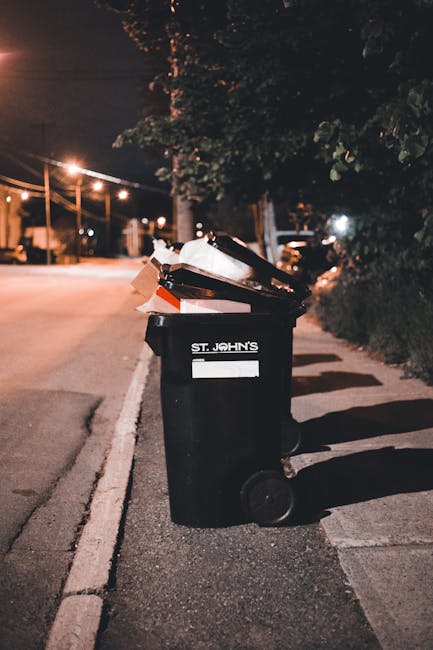 A black wheelie bin positioned on a quiet residential street at night, with the label 'ST. JOHN'S' visible on the front. The bin is filled with various waste materials, including paper, cardboard, and