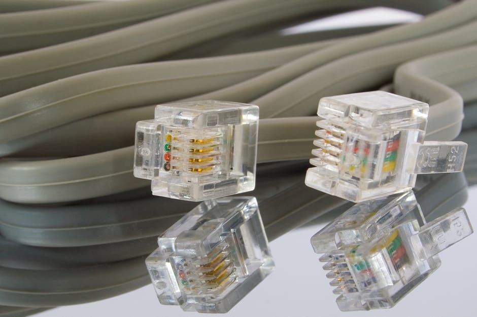 A close-up view of a coiled gray Ethernet cable with four transparent RJ45 connectors resting on a reflective surface. The connectors reveal gold-plated pins and colored wiring inside, with some showi